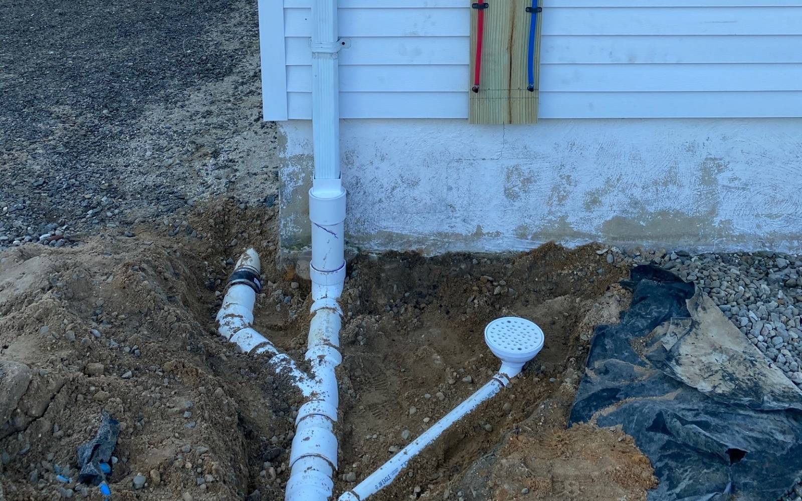 Excavated area showing a section of white plumbing pipes connected to a drain system near the foundation of a house.
