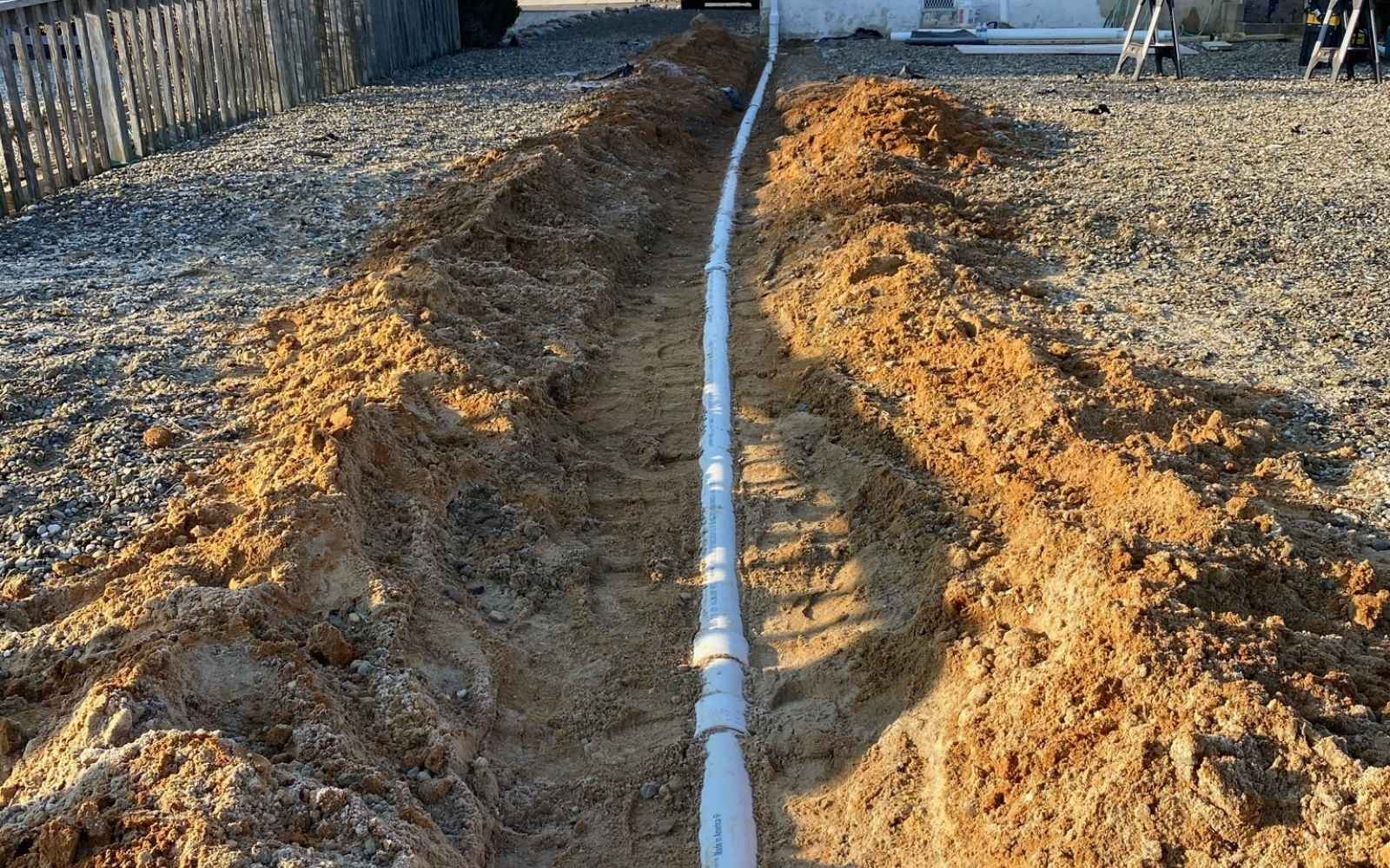 user-inserted A long section of white drainage pipe installed in an excavated trench running alongside the foundation of a building.
