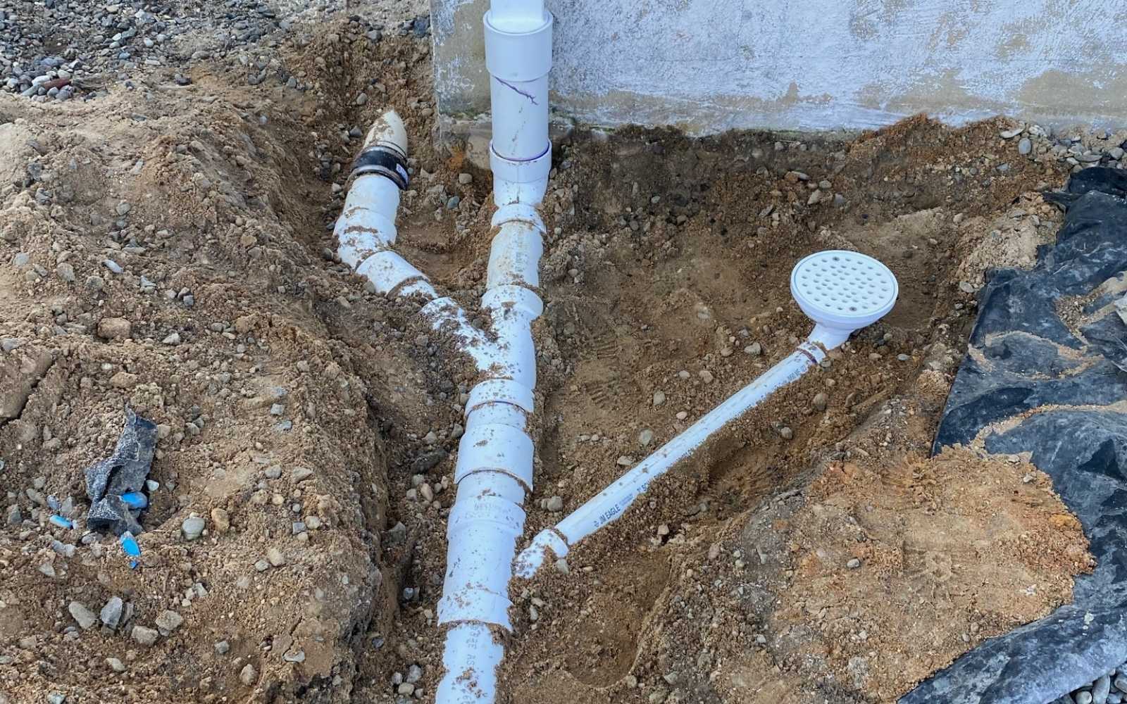 Excavated area showing a section of white plumbing pipes connected to a drain system near the foundation of a house.