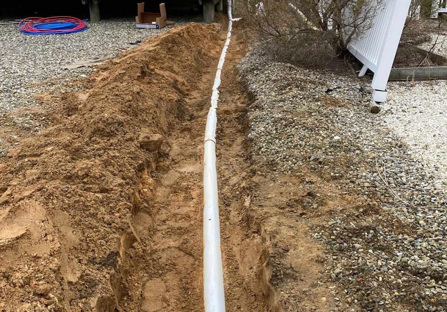 A trench dug in the ground with a white pipe running through it, likely for drainage, outside a home.